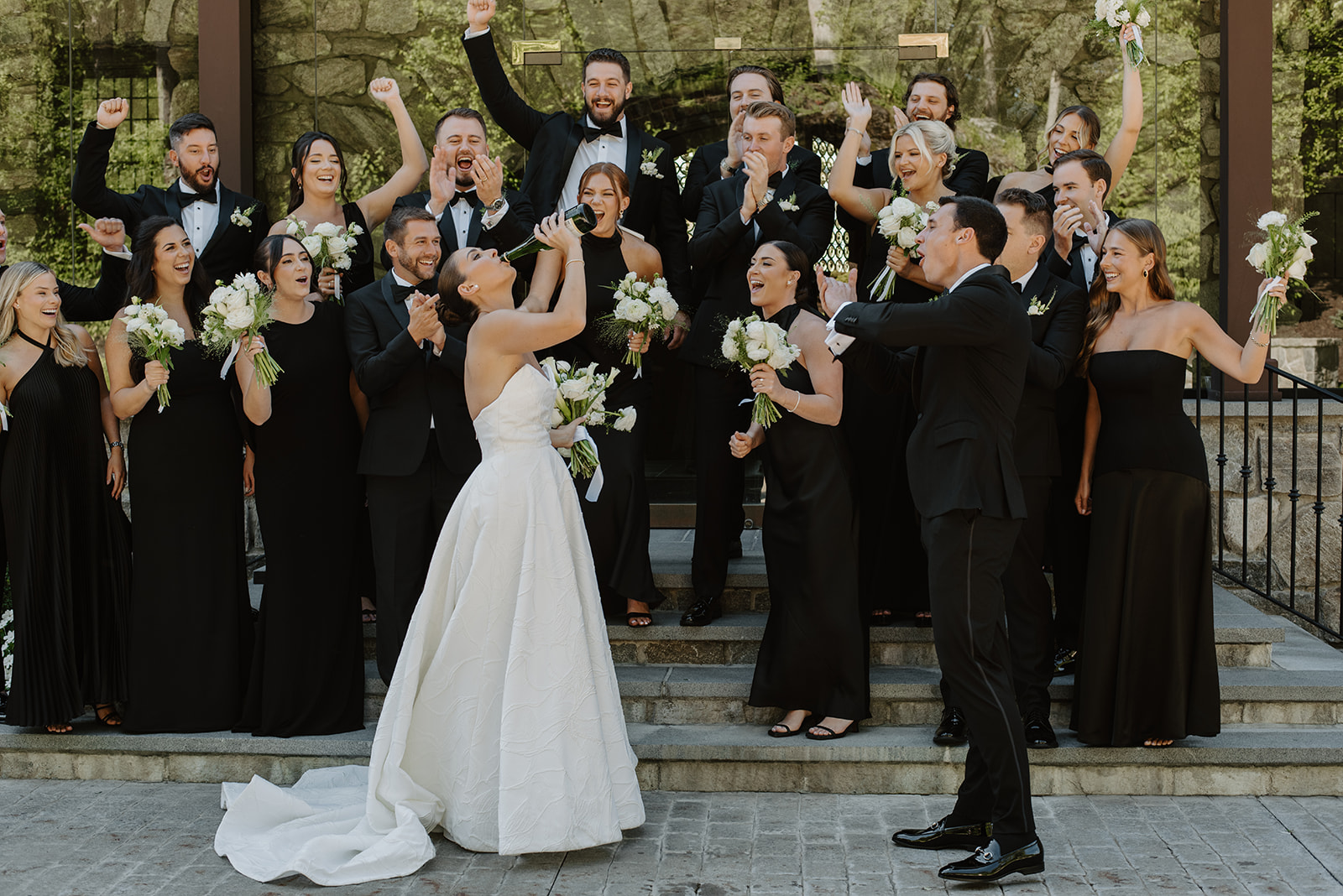 Bride drinking straight from a champagne bottle while the groom and wedding party cheer her on.